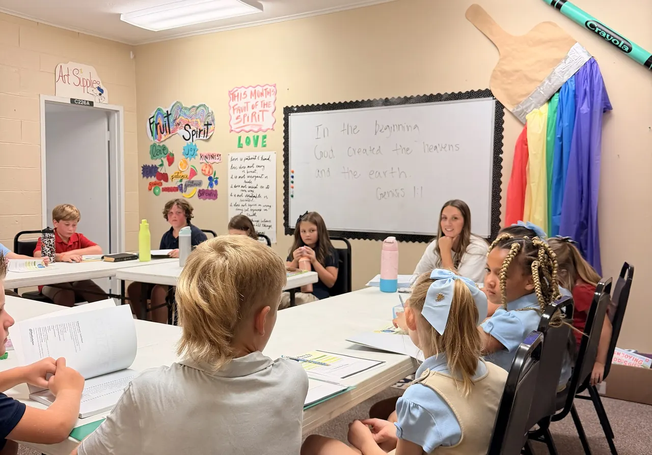 Anchor Christian students gathered around tables for a Bible lesson with their teacher, Genesis 1:1 written on the whiteboard